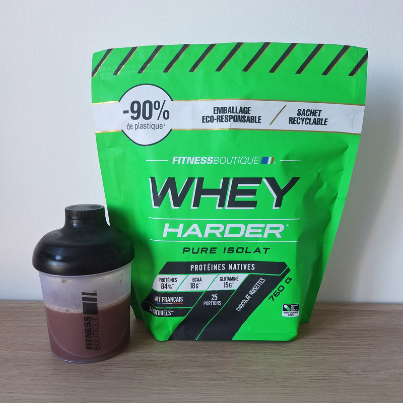 Photo du sachet de la whey Harder saveur chocolat / noisettes et d'un shaker avec 250 ml d'eau sur le bureau de Calvin
